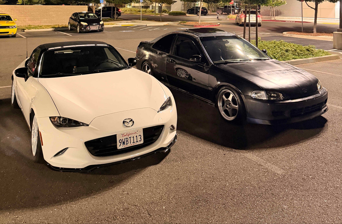 Two cars parked in a nighttime outdoor lot: a white Mazda MX-5 Miata in the foreground and a black sedan behind it, with streetlights, trees, and buildings visible in the background.